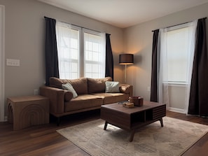 A living room with a brown sofa, a wooden coffee table, and a lamp. There are two windows with curtains, a small side table, and a rug on the floor.