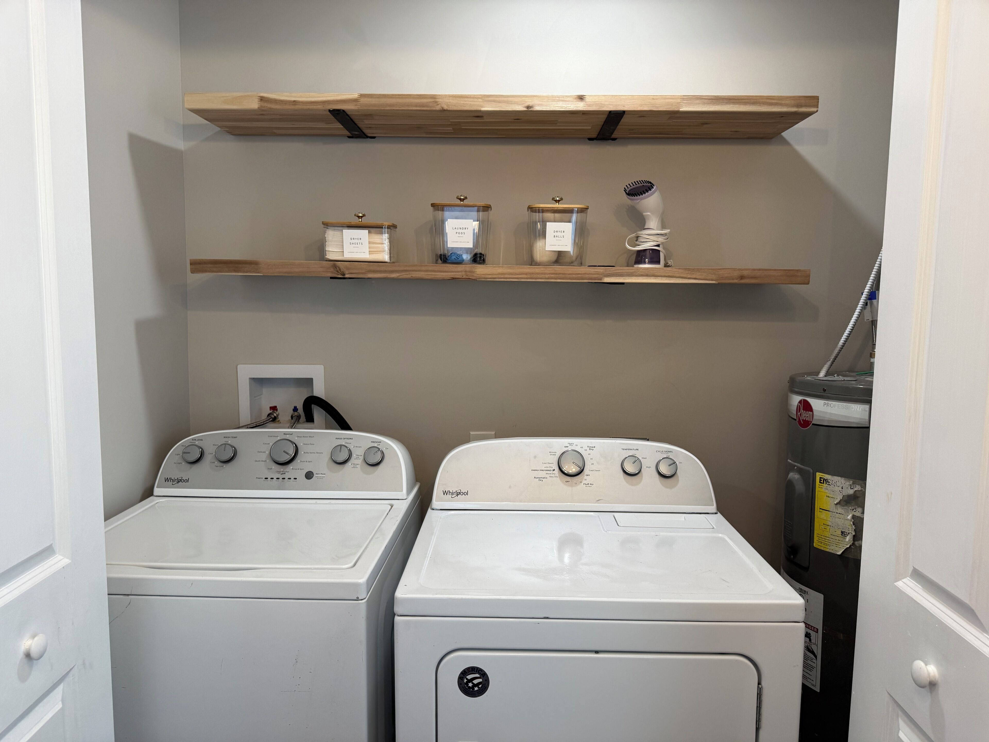 A laundry room with two Whirlpool washers, a shelf above with storage bins, and a professional-grade dryer.