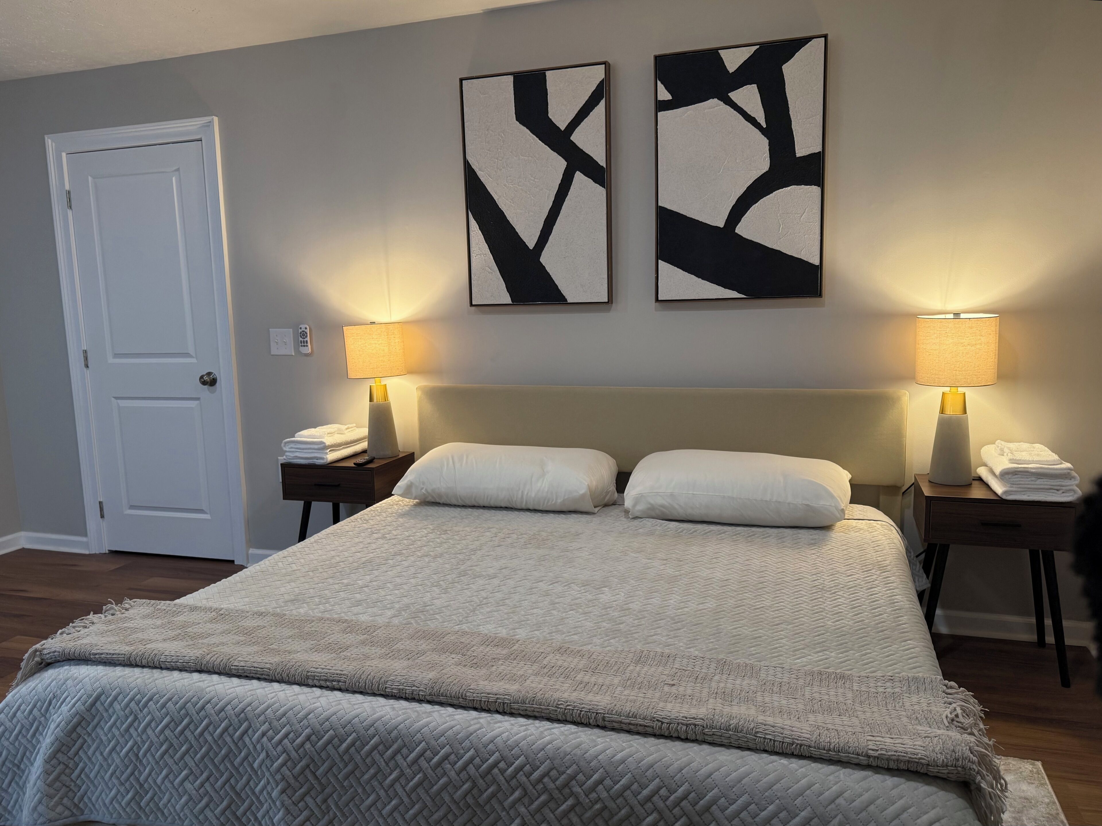 Ein Hotelzimmer mit einem großen Bett, zwei Nachttischen mit Lampen, einer Tür und abstrakter Wandkunst.