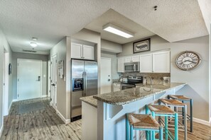 A modern kitchen with a central island, bar stools, stainless steel appliances, and a clock on the wall. - Tides 401 Corner Unit-Pool- Overlooks the Gulf! (Miramar Beach)