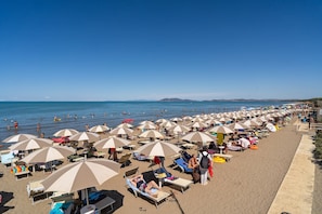 Ein Strand mit vielen Sonnenschirmen und Liegestühlen, ein sandiger Uferstreifen und das Meer im Hintergrund.