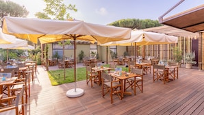 Un patio al aire libre con mesas y sillas de madera, cubierto por sombrillas grandes, y una terraza de madera.