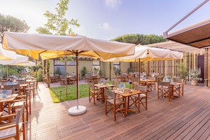 Eine Terrasse im Freien mit Holztischen und -stühlen, beschattet durch große Sonnenschirme, und ein Holzdeck.