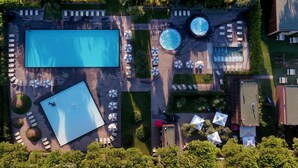 Vista aérea de un complejo turístico con una amplia piscina rectangular, piscinas circulares más pequeñas y numerosos sillones de sol y sombrillas.