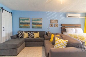 A modern bedroom with a bed, a sofa, and a small table. There are two paintings on the wall and an air conditioning unit above the bed. - Charming studio condo with a coastal comfort (Christiansted)