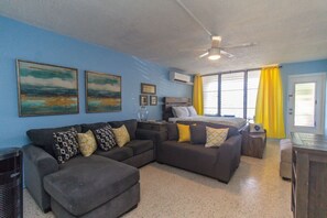 A room with a sofa, a bed, a ceiling fan, and a window with yellow curtains. - Charming studio condo with a coastal comfort (Christiansted)