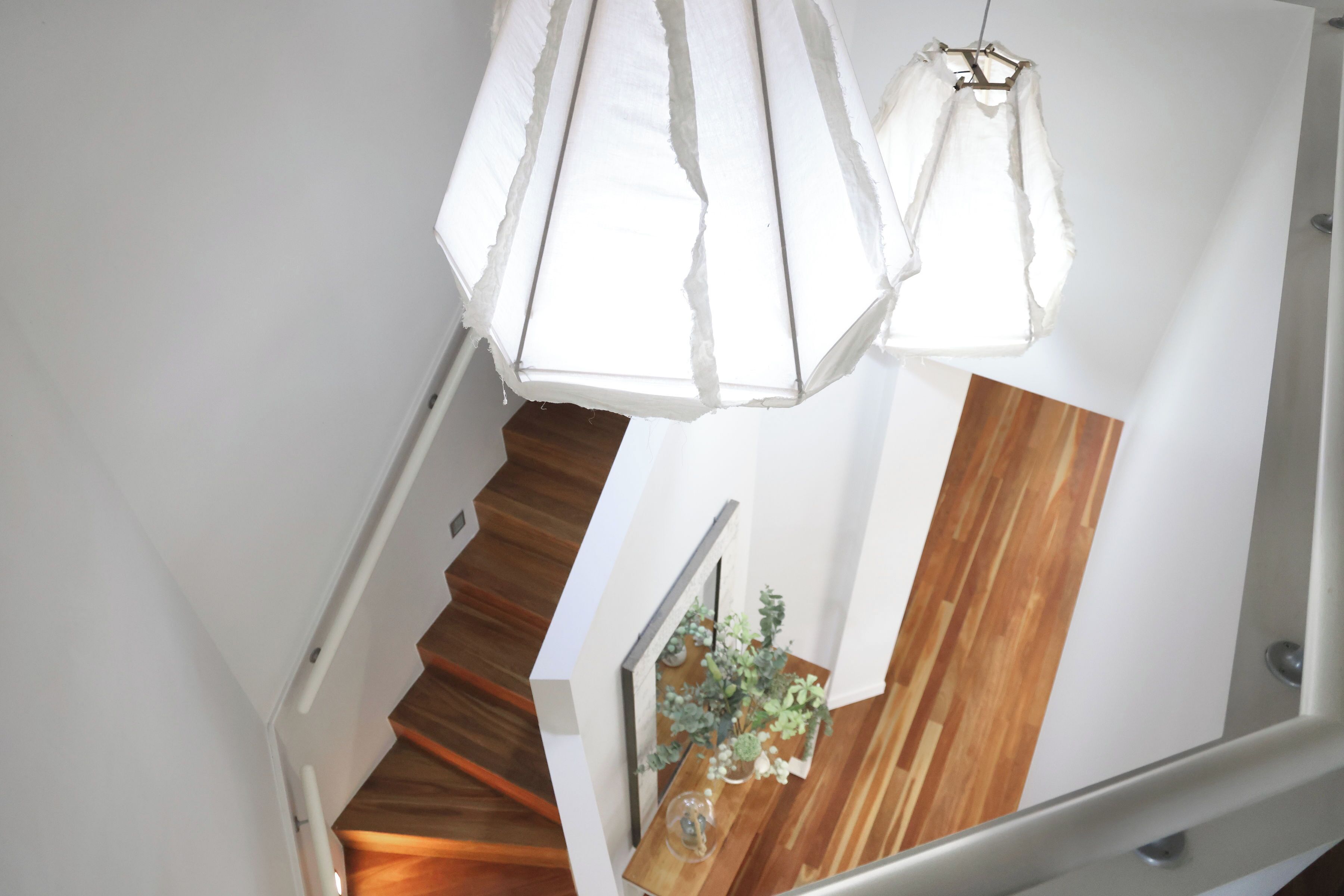 A staircase with wooden steps and a white wall. There is a wooden shelf with plants and a vase. Two white pendant lights hang from the ceiling.