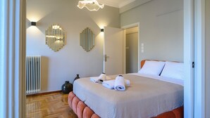 Una habitación de hotel con una cama grande, dos lámparas de pared, un radiador y una puerta que da a otra habitación.