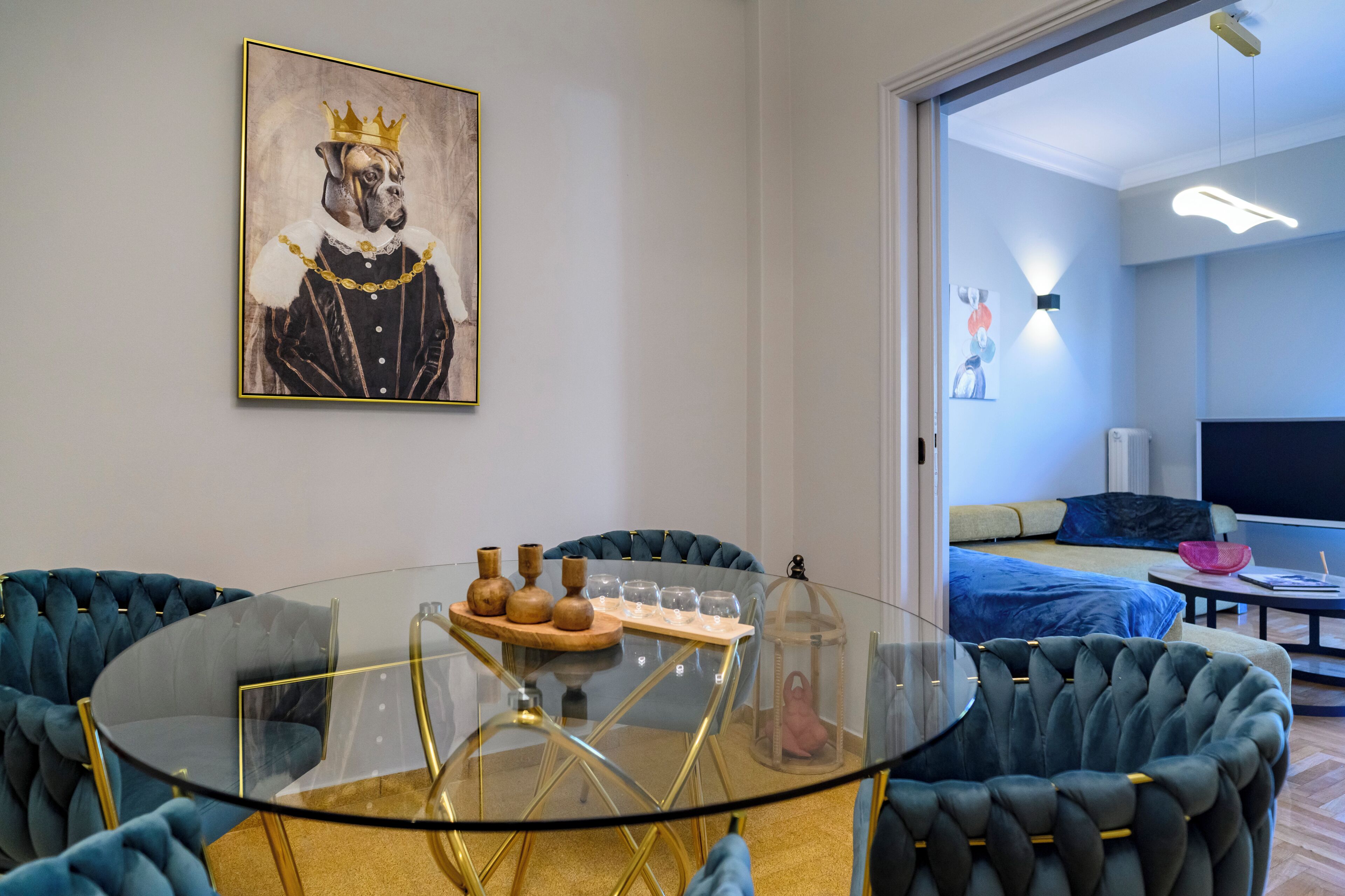 A modern room with a glass table, blue upholstered chairs, a painting of a dog in a crown, and a bed with a blue cover.