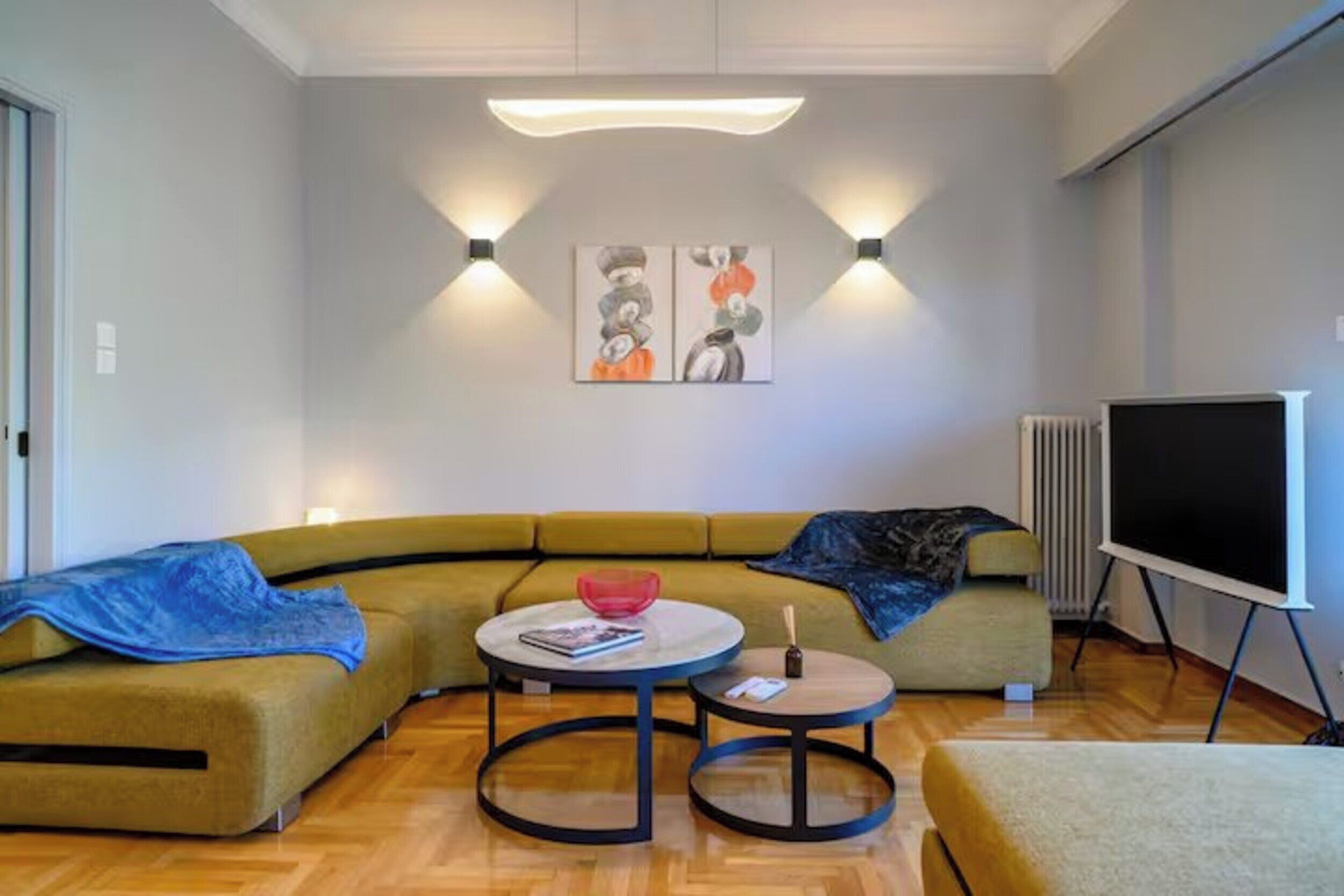 A modern living room with a sectional sofa, two round tables, a flat-screen TV, and wall-mounted lighting.
