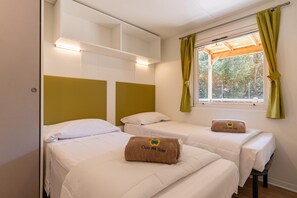 A room with two beds, each with a white bedspread and pillows, and a brown towel with 'Club del Sole' on it. There is a window with green curtains and a shelf above the beds. - Mobilhome im Ferienpark mit Grosser Poolanlage (Castiglione della Pescaia)