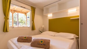 A hotel room with a bed, two towels with the hotel's logo, a window with curtains, and a view of trees outside. - Mobilhome im Ferienpark mit Grosser Poolanlage (Castiglione della Pescaia)