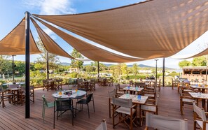 An outdoor dining area with tables and chairs under large shade structures, overlooking a scenic landscape with hills and a body of water. - Mobilhome im Ferienpark mit Grosser Poolanlage (Castiglione della Pescaia)
