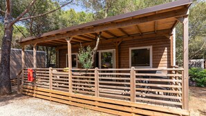 A wooden cabin with a covered porch, a fenced area, and a red object on the ground. - Mobilhome im Ferienpark mit Grosser Poolanlage (Castiglione della Pescaia)