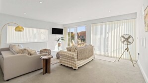 A spacious living room with a sofa, armchair, and a small side table. There is a large window with blinds, a flat-screen TV mounted on the wall, and a standing fan. - Beachside Coastal Retreat with Ocean Views (Pevensey Bay)