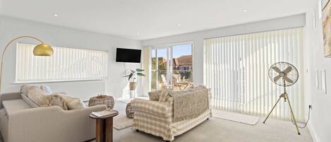 A spacious living room with a sofa, armchair, and a small side table. There is a large window with blinds, a flat-screen TV mounted on the wall, and a standing fan.