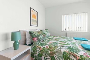 A bedroom with a bed, a nightstand, a lamp, a framed picture on the wall, and a window with blinds.