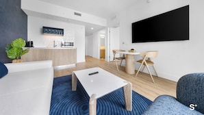 Ein modernes Wohnzimmer mit einem weißen Sofa, einem Couchtisch und einem blauen Teppich. Es gibt eine Küchenzeile mit einer Bar und einem Esstisch mit Stühlen.