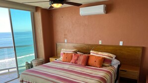 A bedroom with a large bed, a ceiling fan, an air conditioning unit, a nightstand, a wicker chair, and a view of the ocean. - Beachfront Condo + Pool - 2 Bedrooms, 2 king beds, 2 baths, w/ AC - Kite Towers (Nuevo Vallarta)