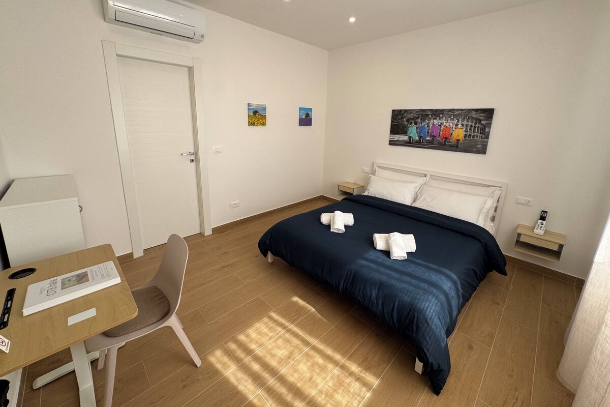 Ein Hotelzimmer mit Bett, Schreibtisch, Stuhl, Klimaanlage, einem Gemälde und einem Nachttisch mit Fernbedienung.