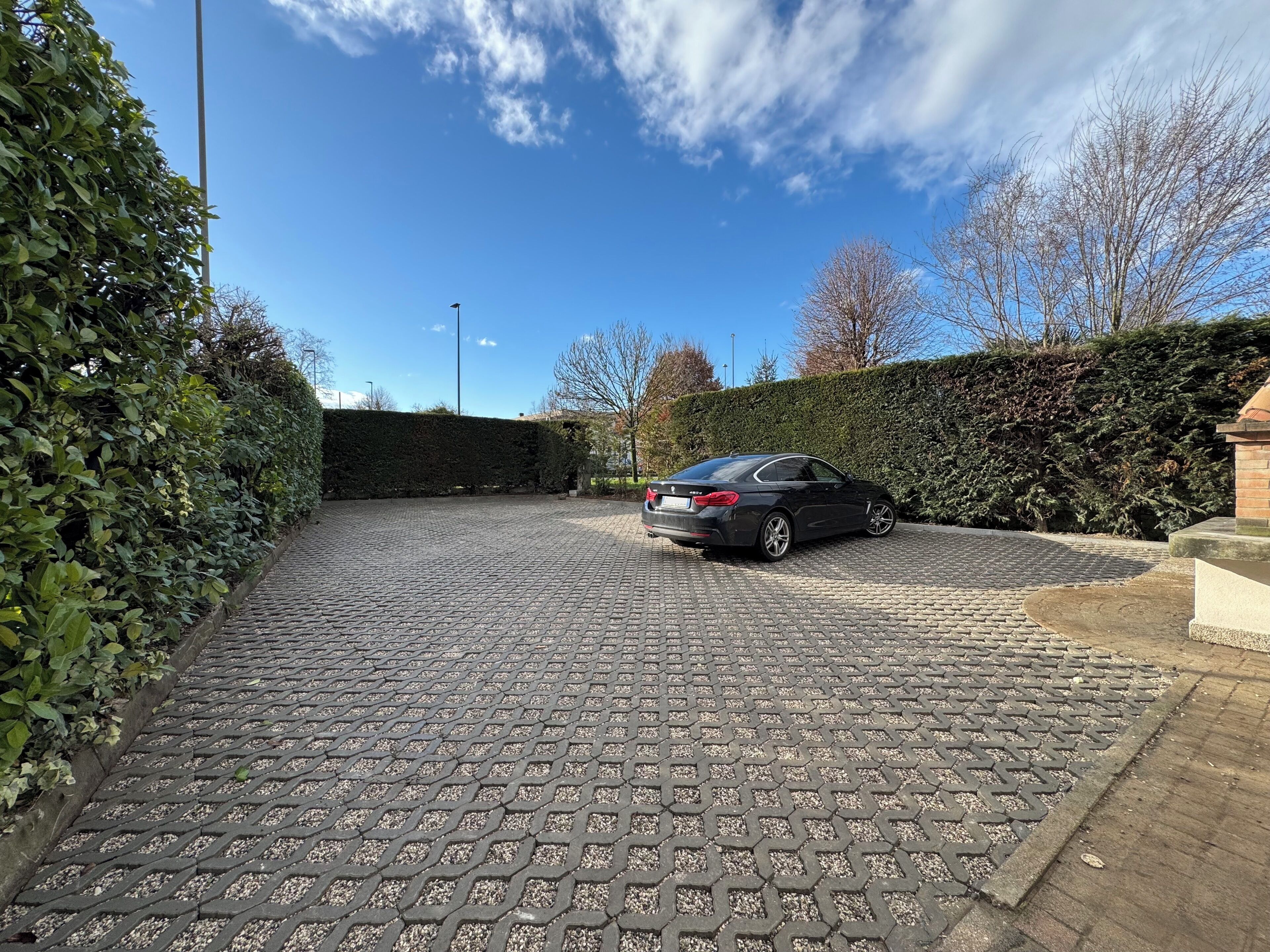 A paved driveway leading to a black car parked in a driveway with a hedge on one side and a brick wall on the other.