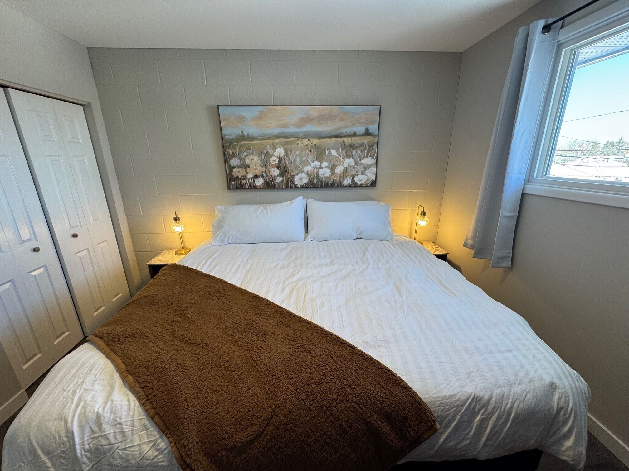 Quarto com cama, duas luminárias de cabeceira, um quadro na parede e janela com cortinas.