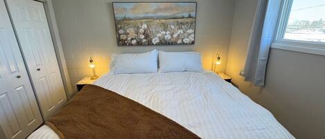 Quarto com cama, duas luminárias de cabeceira, um quadro na parede e janela com cortinas.
