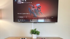 Seinälle asennettu televisio, jossa näkyy peli «Marvel’s Spider-Man 2». Sen alla olevassa puukalusteessa on peliohjain ja kasvi.