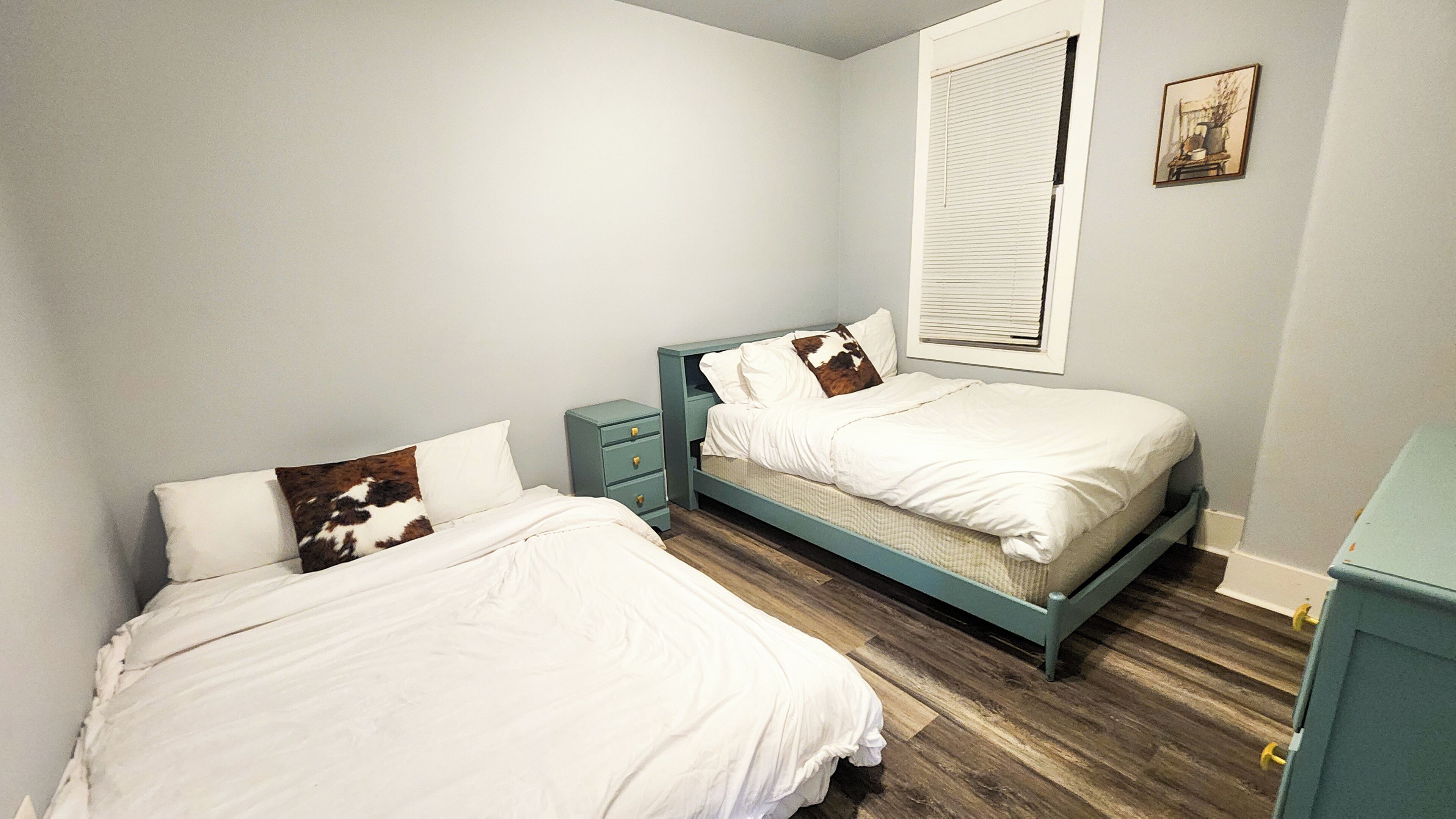 Une chambre avec deux lits simples, une table de chevet, une fenêtre avec des stores et un petit tableau au mur.