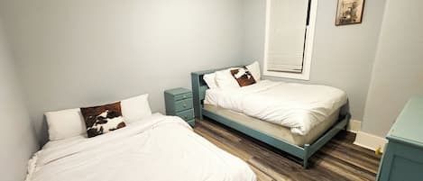 Une chambre avec deux lits simples, une table de chevet, une fenêtre avec des stores et un petit tableau au mur.