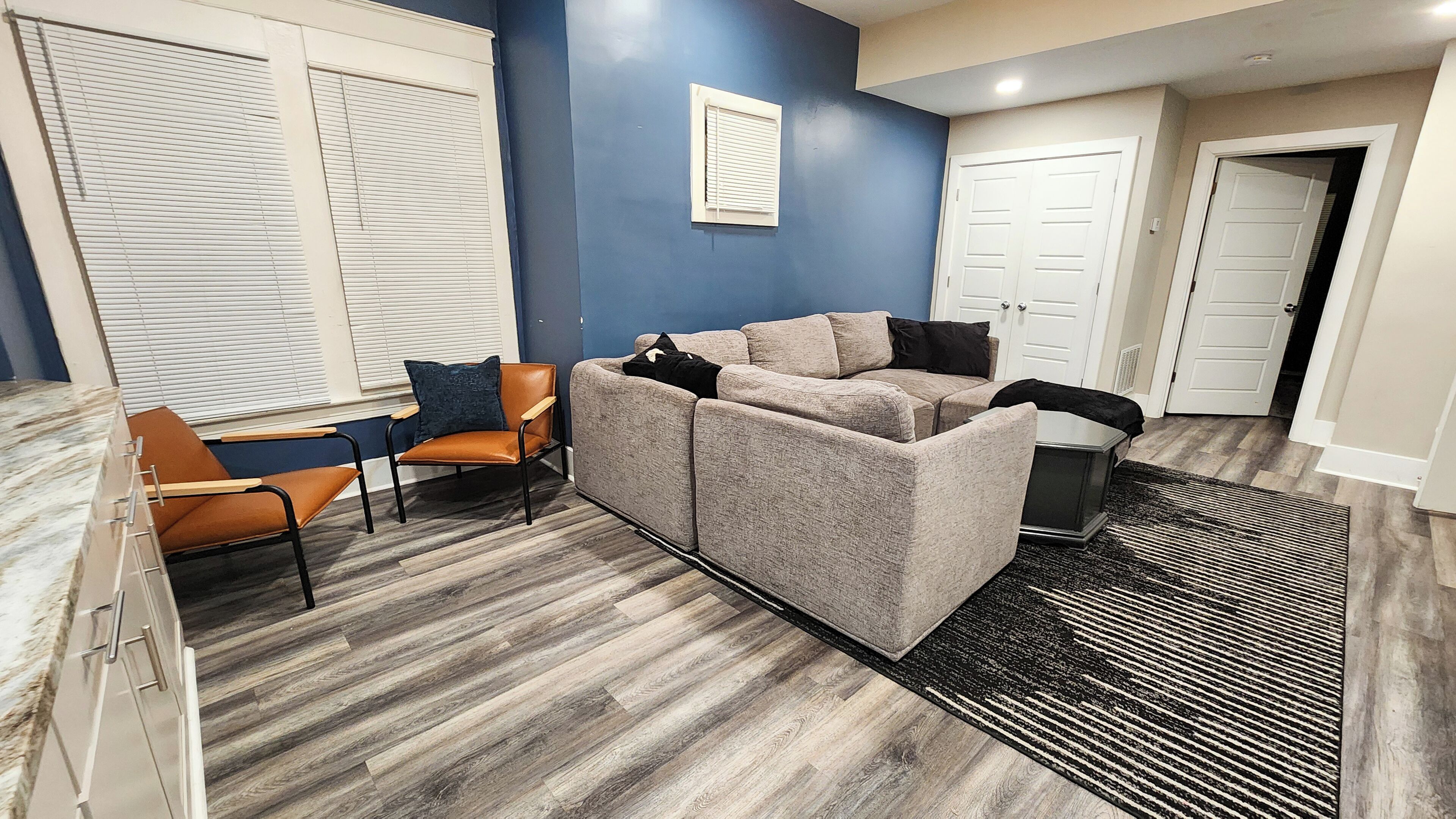 Sala de estar con sofá modular, una silla y una mesita. Hay dos puertas y una ventana con persianas.