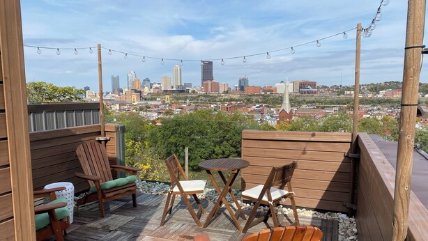 A wooden deck with outdoor furniture, a view of a city skyline, and string lights. - Modern Living with Rooftop Views (Pittsburgh)