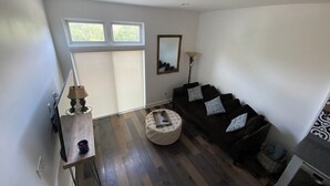 A modern living room with a dark brown sofa, a white ottoman, and a lamp. There are two windows with blinds, a mirror on the wall, and a wooden floor. - Modern Living with Rooftop Views (Pittsburgh)