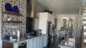 A modern kitchen with a stainless steel refrigerator, a built-in oven, and a dishwasher. There is a wooden dining table with chairs, and a decorative shelf with blue bowls. - Modern Living with Rooftop Views (Pittsburgh)