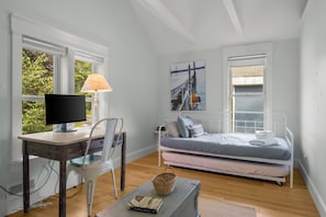 A bedroom with a bed, a desk with a computer, a chair, and a window with a view of greenery.