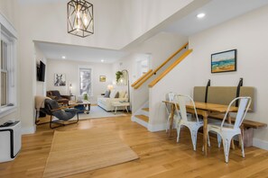A spacious living area with a wooden floor, a dining table with chairs, a sofa, a TV mounted on the wall, and a staircase with wooden railings.