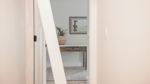 Un couloir avec une porte blanche, un petit bureau orné d’une plante et un tableau encadré accroché au mur.
