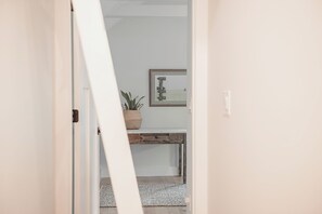 Un pasillo con una puerta blanca, un escritorio pequeño con una planta y un cuadro en la pared.