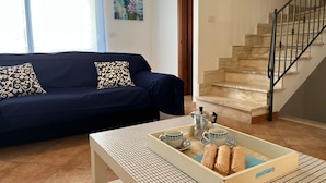 Een woonkamer met een donkerblauwe bank, gestreepte kussens, een witte salontafel met een schaal gebakjes, en een trap met een zwarte balustrade.