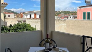 Een balkon met een tafel voor twee, een fles wijn en twee glazen. Het balkon heeft uitzicht op een stadsgezicht met gebouwen en een heldere lucht.