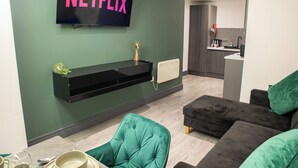Ein modernes Wohnzimmer mit einem Flachbildfernseher, der "NETFLIX" anzeigt, einer schwarzen Unterhaltungseinheit, einem grünen Sessel und einem dunklen Sofa mit grünen Kissen.