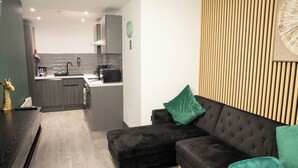 Ein modernes Wohnzimmer mit einem dunkelgrauen Sofa, einer Wand als Holzoptik und einer Küche im Hintergrund.