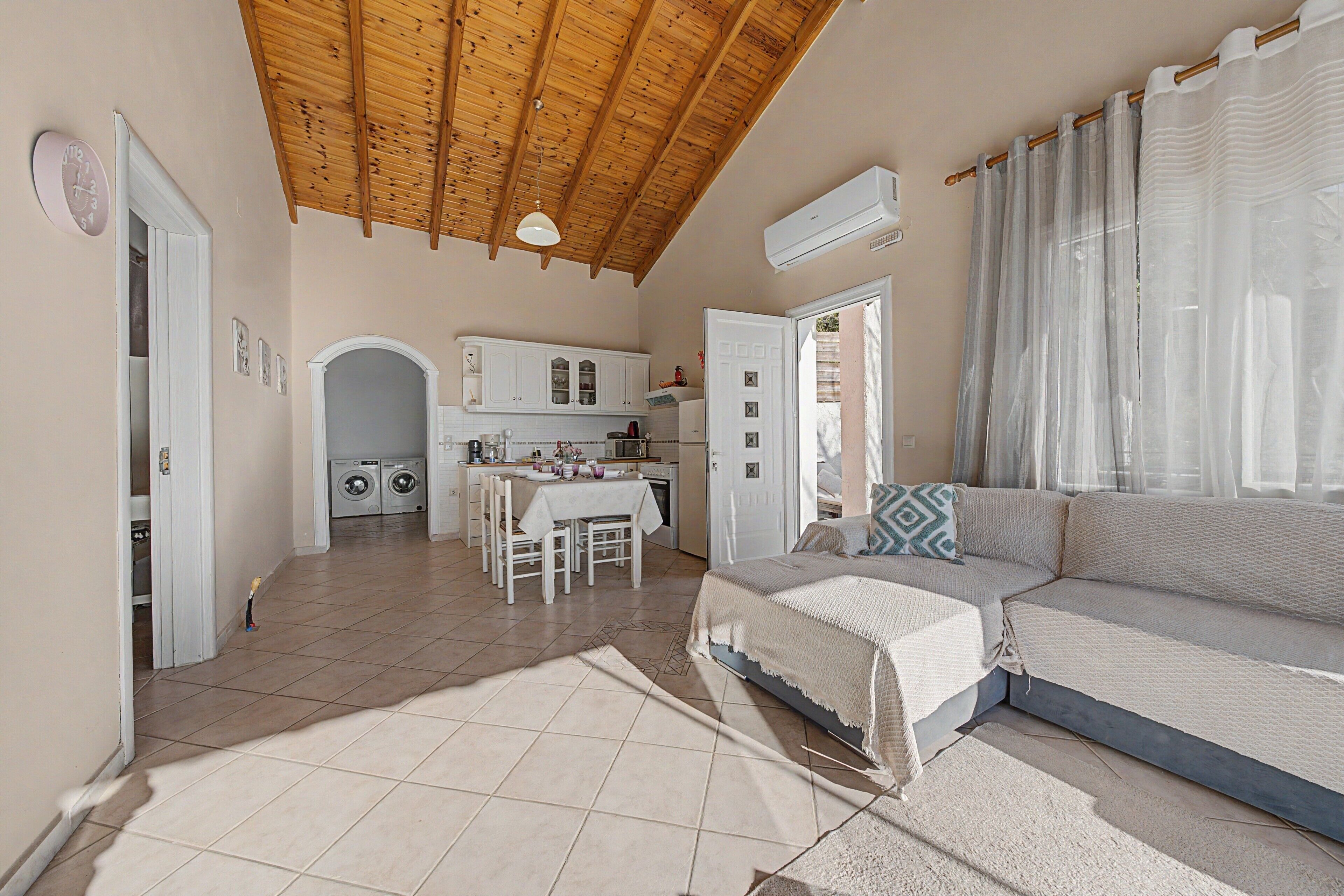 A spacious living area with a high wooden ceiling, a sofa, a dining table, and a kitchen in the background.