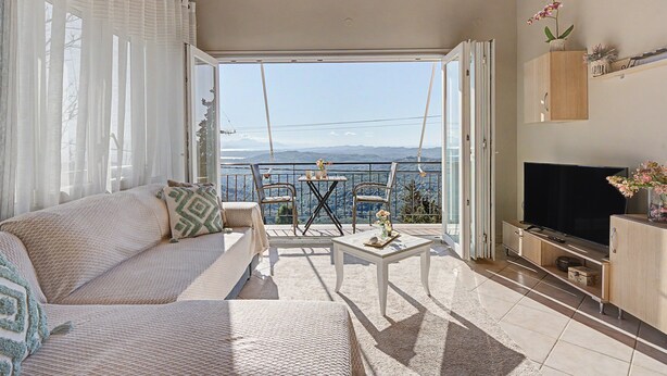 A balcony with a sofa, chairs, a table, a TV, and a view of the mountains. - Dorothea Apartment in Chlomos with Sea View, Wi-Fi and Air Conditioning (Χλομός)
