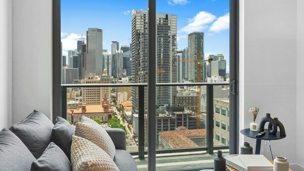 A modern room with a large window, a grey sofa, a small table with a vase and book, and a view of a city skyline. - New 1BR+Den | Rooftop Deck |Free Parking (Miami)
