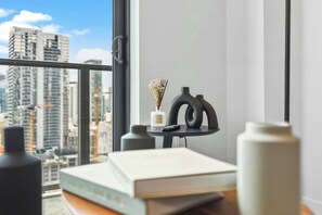 Ein Zimmer mit Blick auf hohe Gebäude, ein kleiner Tisch mit einer Vase und einem Buch sowie ein Fenster mit Griff.