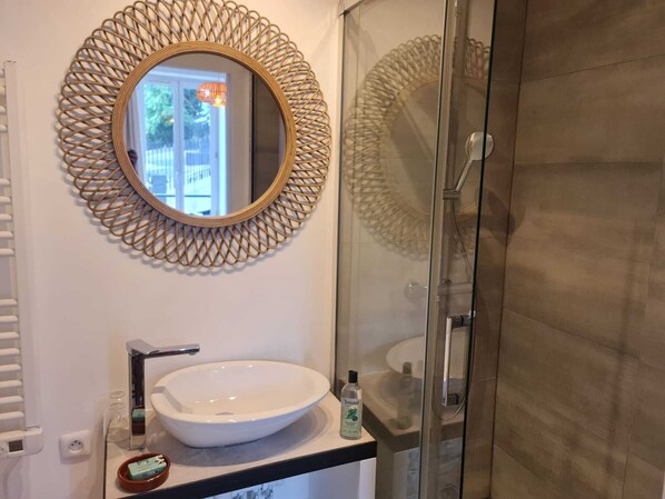 Une salle de bain avec un miroir rond, un lavabo blanc, une cabine de douche en verre et une vue sur l’extérieur.