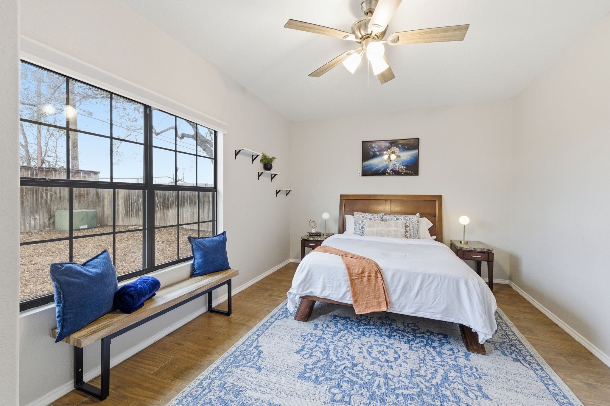 A bedroom with a large window, a bed with a white and orange blanket, a wooden bench with blue cushions, a ceiling fan, and a nightstand with a lamp.