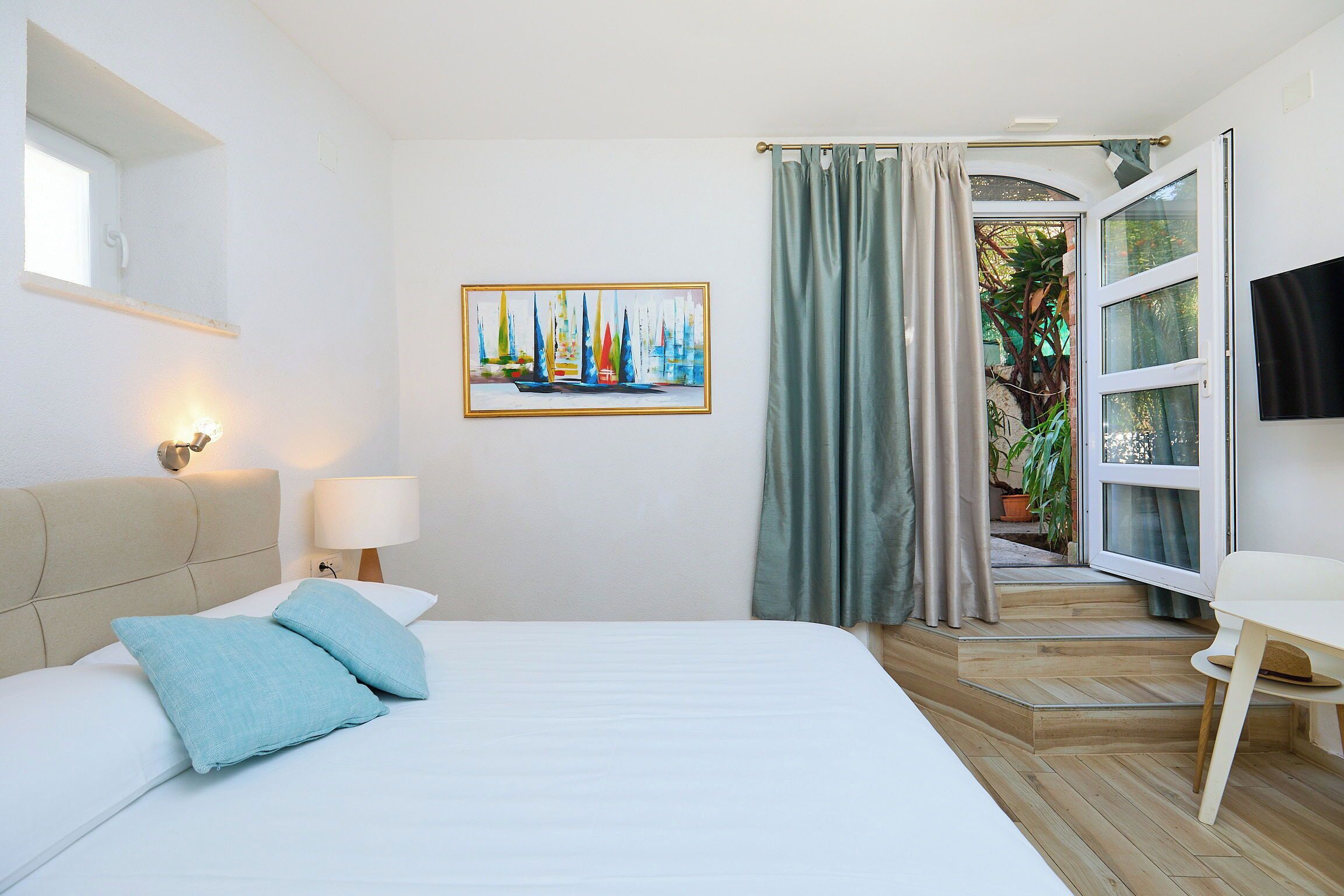 A bedroom with a bed, a nightstand, a lamp, a painting, a window, and a door leading to a balcony.