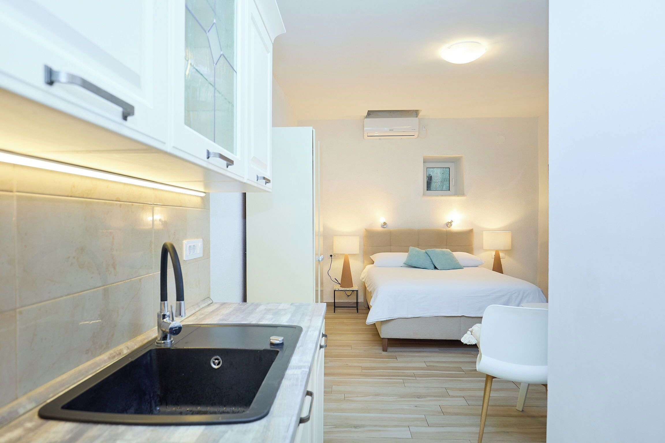 A compact hotel room with a kitchenette, a bed with white linens, a bedside table with a lamp, and a small bedside table with a picture.